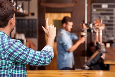 Cheerful man is ordering beer in bar. He is sitting at counter and gesturing to barman. The worker...
