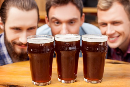Cheerful three men are looking at glasses of beer on table with desire. They are smiling and resting in pub. Focus on lagerの写真素材