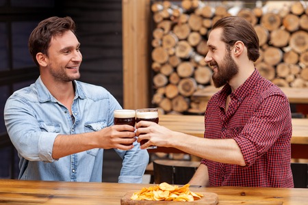 Attractive two young guys are drinking beer in bar. They are sitting and clinking glasses. The...