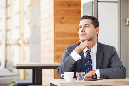 Cheerful young businessman is sitting and thinking seriously in cafeteria outdoors. He is touching his chin and looking forward pensively. The man is drinking coffee. Copy space in left sideの写真素材