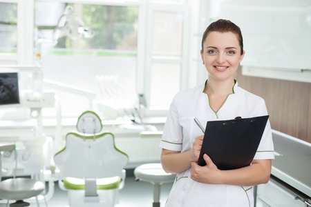 Beautiful young dental doctor is holding document and writing data. She is standing and smiling. The woman is looking forward happily. Copy space in left sideの写真素材