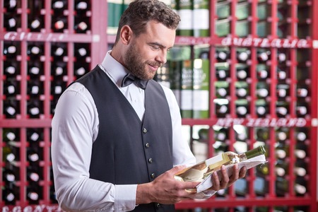 Handsome wine waiter is standing in liquor store. He is holding a bottle of white wine and looking at it with joy. The man is smilingの写真素材