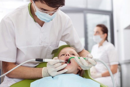 Cheerful young dentist is drilling teeth of child. The man is holding tools and looking at kid with seriousness. Boy is sitting and opening mouth with fear. Female assistant is standing on backgroundの写真素材