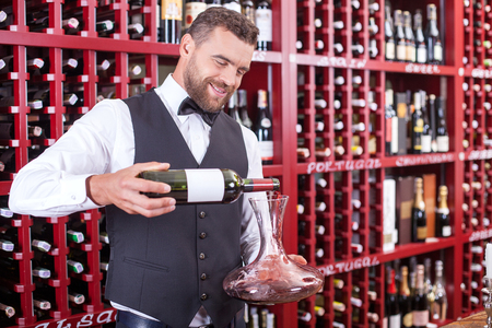 Cheerful male sommelier is holding a bottle of wine. He is pouring it into carafe. The man is looking at drink and smiling. He is standing in cellarの写真素材