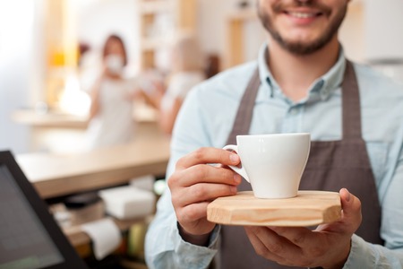 Close up of worker of cafeteria. The man is holding a cup of coffee and giving it with joy. The man is standing and smiling. Two women are drinking espresso and talking on backgroundの写真素材