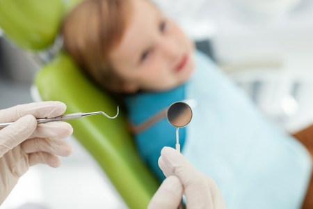 Close up of hands of professional dentist standing near the child. The man is holding tools. The boy is sitting in medical chair with joy. Focus on hook and mirrorの写真素材