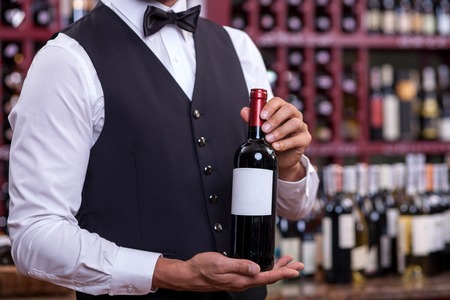 Close up of hands of wine waiter. The man is holding a bottle of wine. He is standing in cellarの写真素材