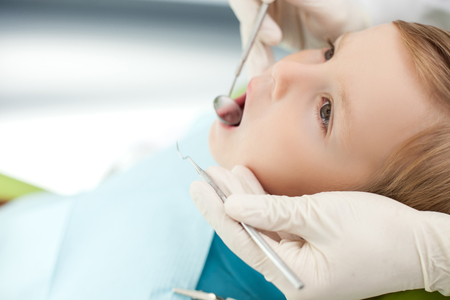 Close up of male hands with glove of dentist checking human teeth. The boy is sitting in chair and looking up with concentration. His mouth is open widelyの写真素材