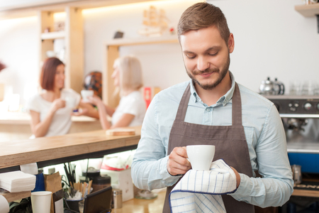 Cheerful young barista is working in coffee house. He is standing and drying a white cup with towel. He is looking at it and smiling. Two beautiful women are drinking coffee and talking on backgroundの写真素材