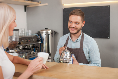 Attractive young barista is standing and working in coffee shop. He is holding a kettle and looking at female client with joy. The woman is holding a cup and smilingの写真素材