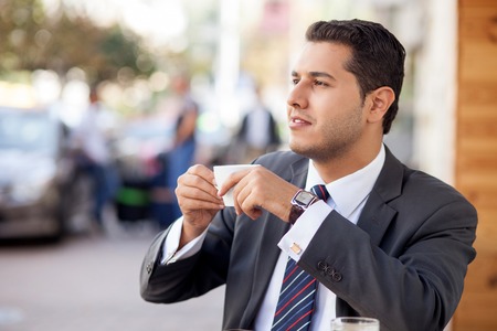 Handsome young man in suit is drinking coffee in cafe. He is sitting outdoors and smiling. The man is viewing city with joy and relaxationの写真素材