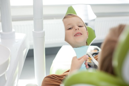 Close up of cheerful boy sitting in dentist chair. He is holding a mirror and looking at his reflection with joy. The kid is satisfied with treatment of his teeth. He is smilingの写真素材