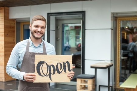 Open. Cheerful male owner of coffee shop is standing outdoors. He is holding placard and inviting everybody to come in. The man is looking at camera and smilingの写真素材