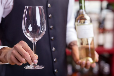 Close up of hands of wine waiter standing in liquor store. The man is holding a bottle of white wine and glass. Focus on wineglassの写真素材