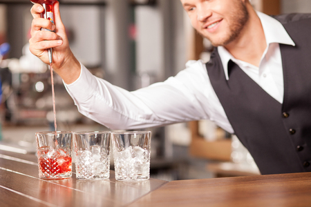 Close up of attractive barman making cocktails in pub. He is pouring drink from bottle into glass. The man is looking at it and smilingの写真素材
