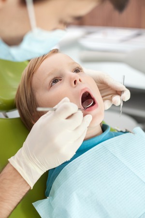 Close up of arms with gloves of experienced dental doctor. The man is examining teeth of kid. The boy is sitting in dental chair and looking up with fear. His mouth is wide openの写真素材
