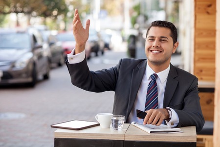 Attractive businessman is gesturing and calling for waiter. He is sitting at table in cafeteria outdoors. The man is looking forward and smiling. He is holding mobile phoneの写真素材