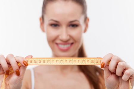 Attractive healthy girl is holding tape-measure and showing numbers to camera. She is standing and...