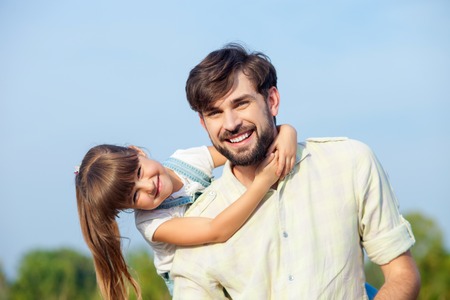 Attractive young man and his daughter are resting in the nature. The father is standing and carrying kid on his back. The girl is embracing him. They are looking forward and smilingの写真素材