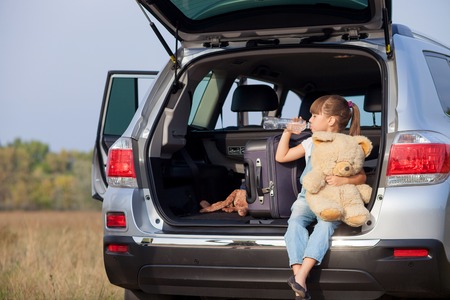Pretty girl is sitting on open car boot in the nature. She is holding bottle and drinking water. Her eyes are closed with pleasure. The kid is holding teddy bearの写真素材