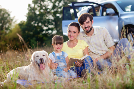Pretty family is making journey in the nature. They are sitting on grass near car and smiling. The mother and father are embracing their daughter. The dog is lying near themの写真素材