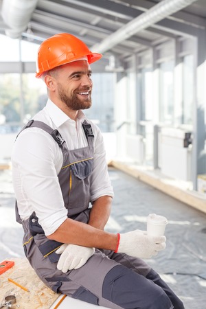 Attractive young builder is drinking coffee on break. He is sitting and smiling. The man is orange helmet is looking forward with happinessの写真素材