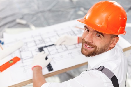 Attractive young architect is projecting building. He is sitting at desk near blueprint. The man in...