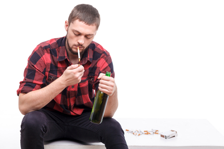 Young bearded man is sitting and smoking. He is lighting cigarette with concentration and drinking bier. Isolated and copy space in right sideの写真素材