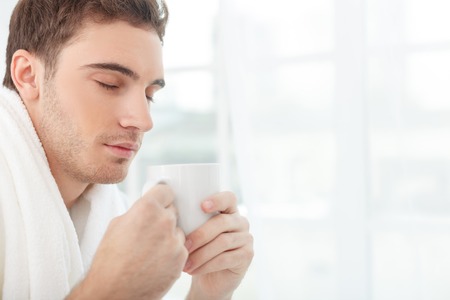 Attractive young man is drinking coffee in the morning. His eyes are closed with pleasure. The man is sitting and relaxing in his bedroom. Copy space in right sideの写真素材