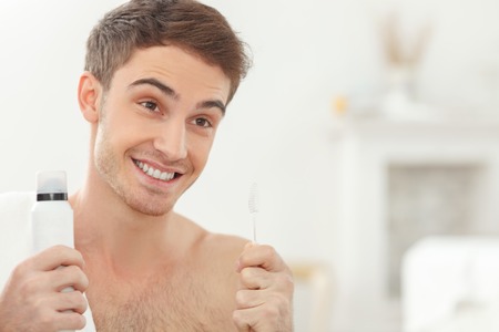 Attractive young man is cleaning his teeth in bathroom. He is standing with a white towel on his neck. The man is holding a tooth-paste and tooth-brush. He is smilingの写真素材