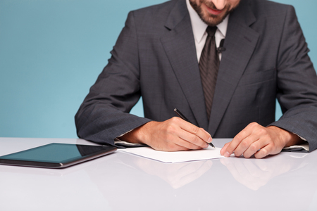 Close up of tv reporter sitting at the desk near a tablet. The man is writing down data into documents. He is smilingの写真素材