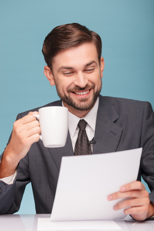 Attractive young tv newscaster is reading documents at studio. He is sitting at desk and drinking a cup of coffee. The man is smiling. Isolated on blue backgroundの写真素材