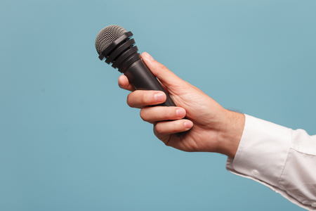 Close up of hand of young reporter interviewing someone. The man is holding a microphone. Isolated on blue background. Copy space in left sideの写真素材