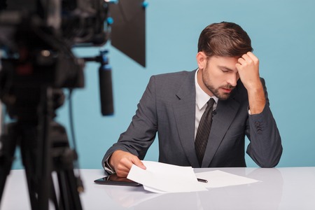 Attractive young tv reporter feels pain in head. He is sitting at desk in studio and touching his forehead with frustration. His tired eyes are closed. Focus on camera and isolated on blue backgroundの写真素材
