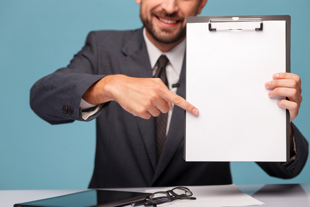 Close up of hands of male tv newscaster showing a folder of white empty paper to the camera. He is pointing finger at it and smiling. The man is sitting at the desk near the tabletの写真素材