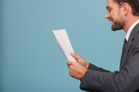Handsome businessman is standing in profile. He is holding papers and reading. He is smiling. Isolated on blue background and copy space in left sideの写真素材