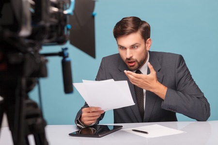 Handsome young tv newscaster is reading papers with shock. He is sitting at the desk in front of the camera. Isolated on blue backgroundの写真素材
