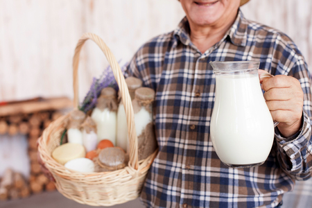 Close up of old farmer showing a jug of milk. He is standing and smiling. The man is holding a basket of dairy productsの写真素材