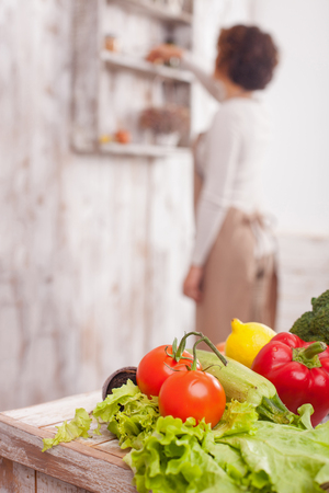 Pretty young housewife is cooking in the kitchen. She is taking the ingredient from the shelf and standing in apron. Focus on the variation of fruits and vegetables on the tableの写真素材