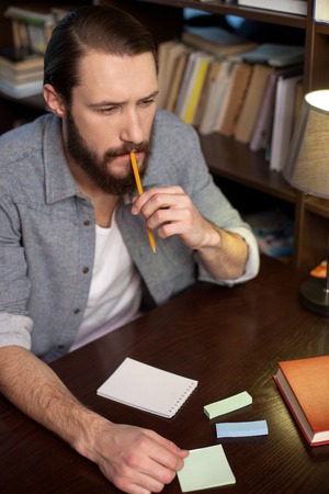 Attractive young student is learning at the library. He is looking forward pensively and touching a pencil to his chin. The man is sitting at the desk neat a bookの写真素材
