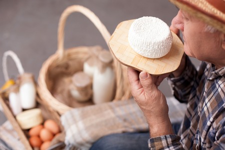Close up of old man holding cheese and smelling it with pleasure. The farmer is sitting near the baskets of dairy products and eggsの写真素材