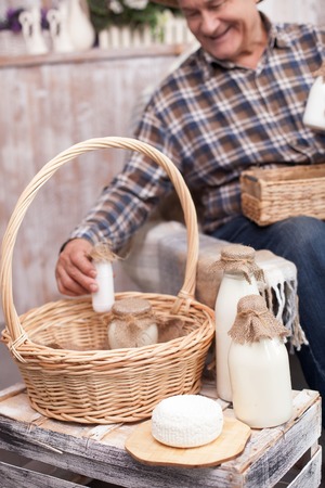 Experienced mature man is taking a bottle of milk from one basket and laying it into another. The man sitting in chair and smilingの写真素材