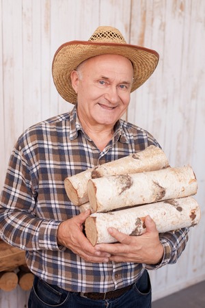 Cheerful old man is holding firewood. He is standing and smiling. The farmer is looking at camera happily. He is wearing a hatの写真素材