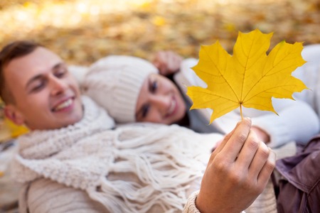 Attractive young loving couple is lying on the blanket in the autumn park. They are embracing and smiling. The man is holding a yellow leaf and looking at it with interest. Focus on leafの写真素材