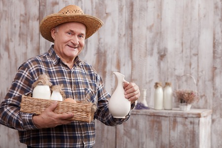 Skillful old farmer is holding a milk jug. He is carrying a basket with eggs and bottles of milk. The man is a straw hat is smiling. He is standing and looking at camera happilyの写真素材