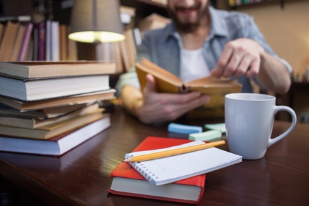 Close up of cheerful young student reading a book at the library. The man is sitting at the desk and smiling. Focus on the cup of tea and books on the tableの写真素材