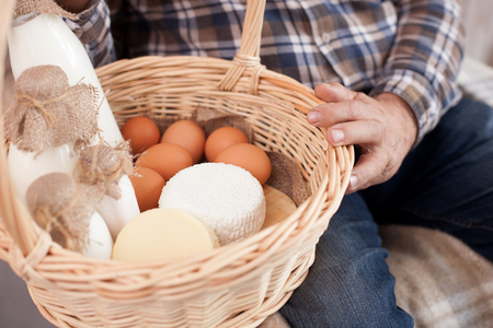 Close up of hands of old farmer sitting in the chair. The man is holding a basket of milk, cheese and eggsの写真素材
