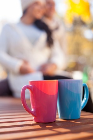 Cheerful young loving couple is dating in the autumn park. They are sitting on bench and embracing. The man and woman are smiling. Focus on two cups of hot coffeeの写真素材