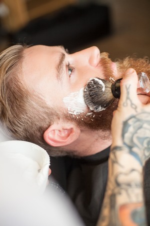 Close up of hands of a barber applying shaving foam on male beard. He is standing and holding a brush. The attractive young hipster man is sitting with relaxationの写真素材