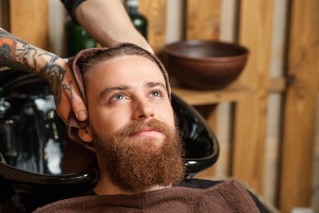 Close up of hands of hairdresser drying male hair with a towel after washing. The young bearded man is leaning his head on the sink. He is smiling with relaxationの写真素材
